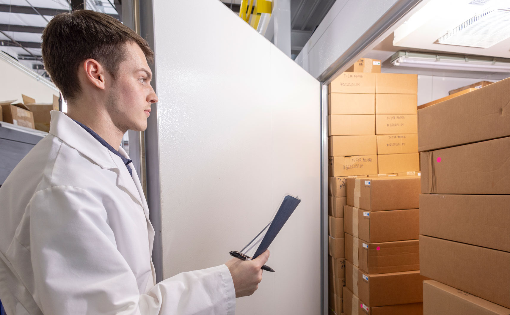 Production technician inspecting boxed products inside an environmental test chamber, documenting conditions during packaging stability and compliance testing.